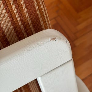 May include: Close-up of a white wooden chair arm with a brown and white striped fabric seat. The paint on the arm is chipped and worn, showing the wood underneath.