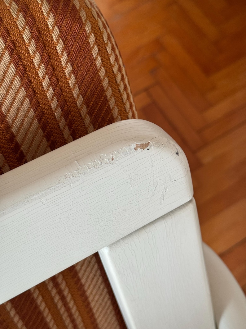 May include: Close-up of a white wooden chair arm with a brown and white striped fabric seat. The paint on the arm is chipped and worn, showing the wood underneath.