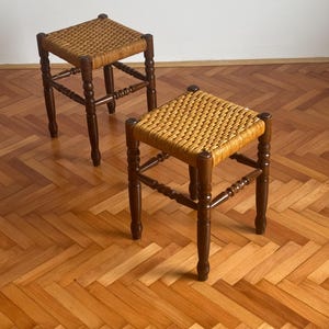 Puede incluir: Dos taburetes de madera con asientos de paja trenzada. Los taburetes tienen un acabado marrón oscuro y están colocados sobre un suelo de madera.