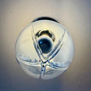 May include: A clear glass globe light fitting with a chrome base. The globe features a distinctive, textured design and a central dark blue element. The light is on, illuminating the interior.
