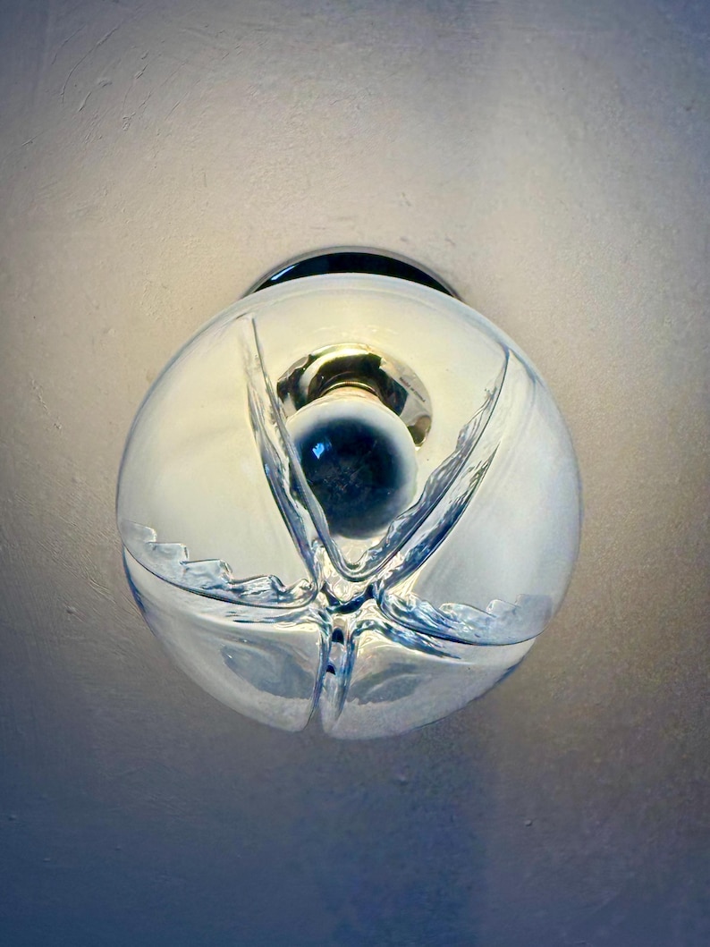 May include: A clear glass globe light fitting with a chrome base. The globe features a distinctive, textured design and a central dark blue element. The light is on, illuminating the interior.
