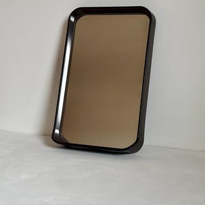 Puede incluir: Un espejo rectangular con esquinas redondeadas, con un marco marrón oscuro y una superficie reflectante con tinte bronce. El espejo está apoyado contra una pared blanca, mostrando un diseño minimalista.