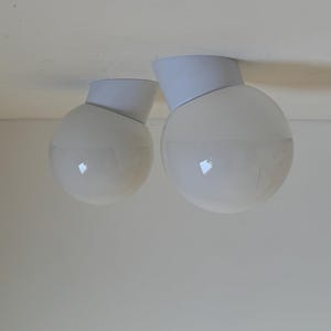 May include: Two white globe-shaped ceiling lights with a modern design. Each light has a white, angled base and a spherical, frosted glass shade. The lights are mounted on a white ceiling, creating a clean, minimalist aesthetic.