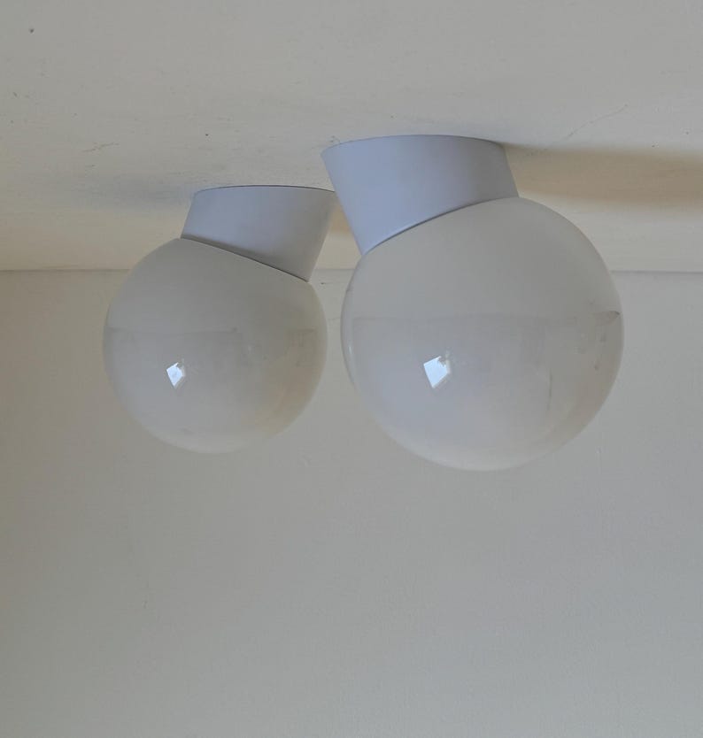 May include: Two white globe-shaped ceiling lights with a modern design. Each light has a white, angled base and a spherical, frosted glass shade. The lights are mounted on a white ceiling, creating a clean, minimalist aesthetic.