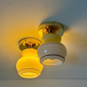 May include: Two vintage ceiling light fixtures. Each fixture has a yellow glass shade with a white base and a gold-colored metal mounting plate. The lights are turned on, emitting a warm, yellow glow. The fixtures are mounted on a white ceiling.