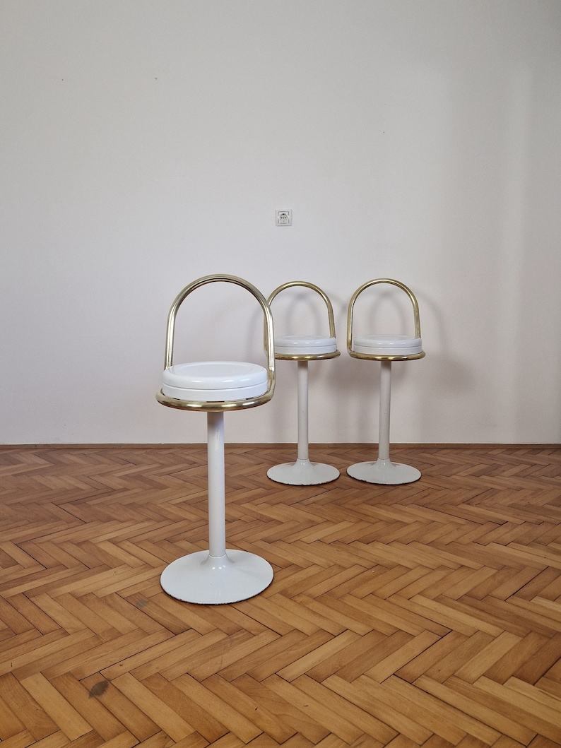 May include: Three white and gold bar stools with a round seat and a curved back. The stools are on a wood floor.