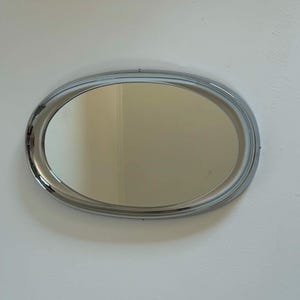 Könnte beinhalten: Ein ovaler Wandspiegel mit einem polierten, reflektierenden Chromrahmen. Der Spiegel verfügt über eine glatte, durchgehende Metallumrandung, die die zentrale Glasscheibe umschließt und ein klares, schlichtes Design für jede Innenwand bietet.
