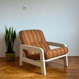 May include: A white armchair with a brown and orange striped fabric upholstery. The chair has a simple design with a wide armrest and a slightly curved back. The chair is sitting on a wooden floor.