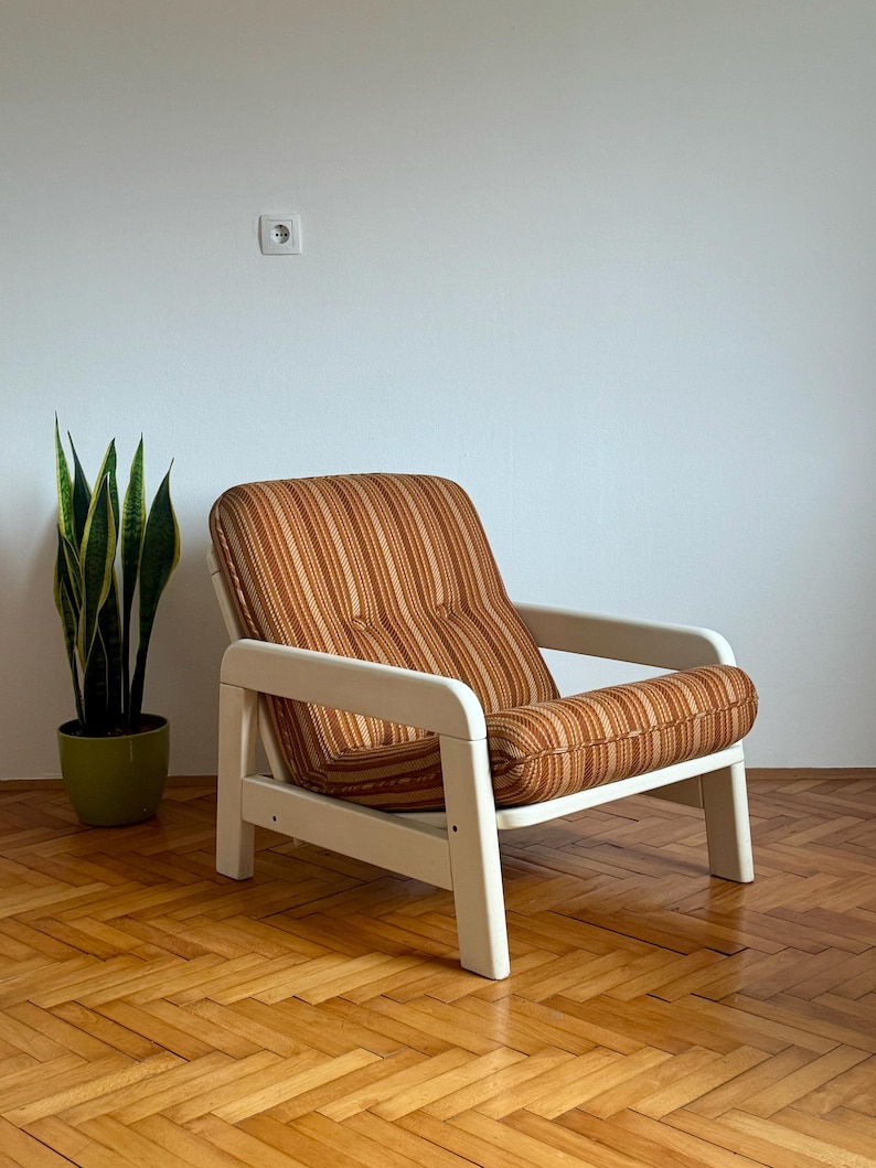 May include: A white armchair with a brown and orange striped fabric upholstery. The chair has a simple design with a wide armrest and a slightly curved back. The chair is sitting on a wooden floor.