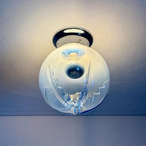 May include: A contemporary ceiling light. It features a spherical, frosted glass shade with a distinctive wave design and blue detailing. The light is mounted on a chrome base and contains a visible light bulb.