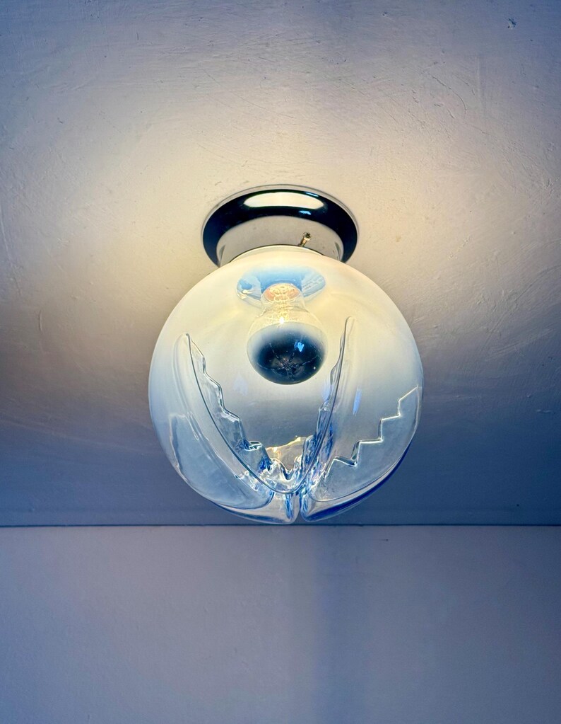 May include: A contemporary ceiling light. It features a spherical, frosted glass shade with a distinctive wave design and blue detailing. The light is mounted on a chrome base and contains a visible light bulb.