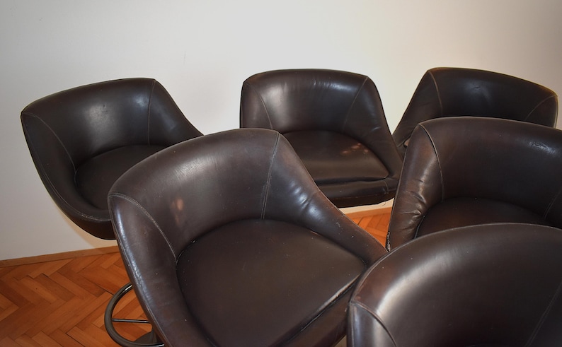 Op de afbeelding: Vijf bruine leren draaistoelen met chromen onderstellen. De stoelen zijn in een halve cirkel gerangschikt.