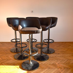 Op de afbeelding: Vijf zwarte barstoelen met bruine leren zittingen en chromen poten. De stoelen zijn in een halve cirkel op een houten vloer geplaatst.