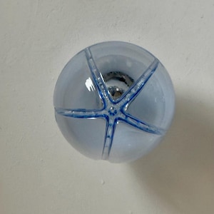 May include: A round, pale blue glass knob featuring a starfish design in a darker blue. The knob is transparent, revealing the hardware within. This decorative knob is suitable for cabinets, drawers, or furniture.