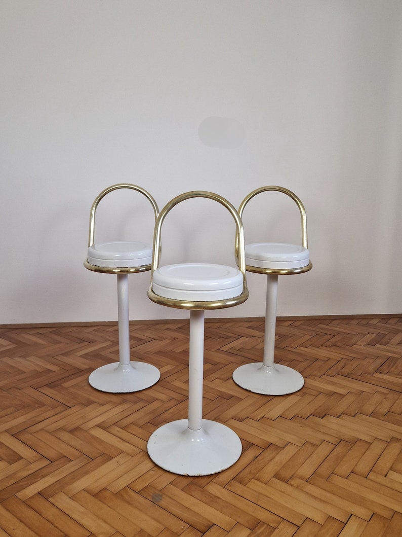 May include: Three white bar stools with gold metal frames. The stools have a round seat and a tall, slender base.