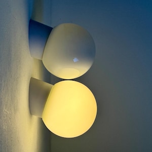 May include: Two modern wall sconces with a spherical, frosted glass shade. The sconces have a white base and a light blue angled support. One is illuminated with a warm yellow glow, the other is unlit.