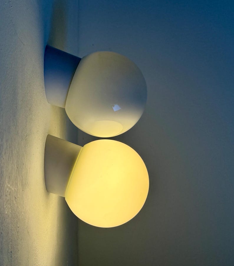 May include: Two modern wall sconces with a spherical, frosted glass shade. The sconces have a white base and a light blue angled support. One is illuminated with a warm yellow glow, the other is unlit.