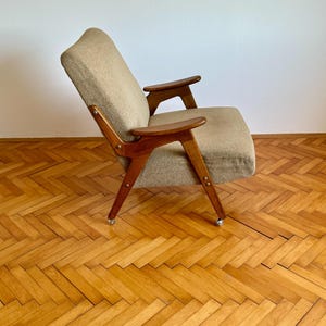 May include: A mid-century modern armchair with a beige fabric upholstery and wooden frame. The chair has a unique design with angled arms and legs. The chair is on a wooden floor with a herringbone pattern.