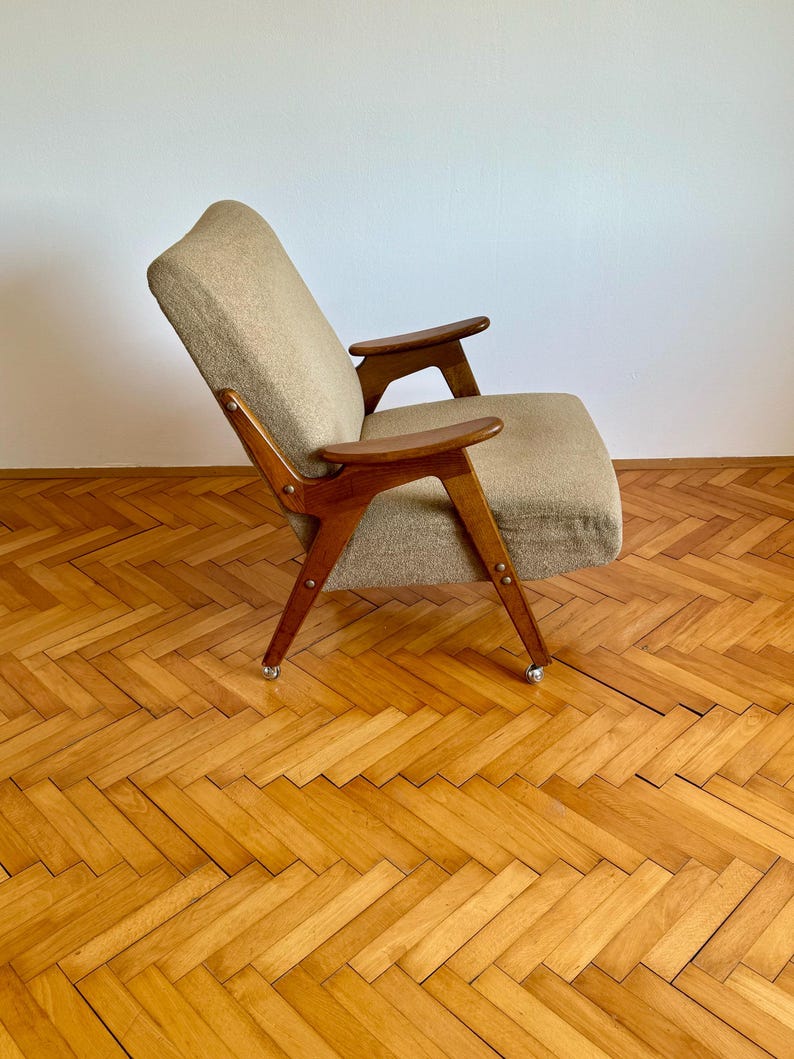 May include: A mid-century modern armchair with a beige fabric upholstery and wooden frame. The chair has a unique design with angled arms and legs. The chair is on a wooden floor with a herringbone pattern.