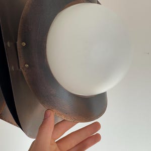 Puede incluir: Una l&aacute;mpara colgante de metal marr&oacute;n con una esfera de vidrio blanco. La l&aacute;mpara tiene un aspecto r&uacute;stico y envejecido.