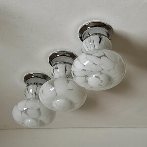 May include: Three flush-mount ceiling lights with chrome bases and white glass globes. The globes have a speckled pattern of white and black. The lights are mounted on a white ceiling.