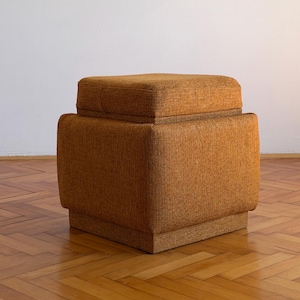 May include: A square, mustard-yellow ottoman upholstered in a textured, woven fabric. The piece features a distinct two-tiered design with a slightly smaller, cushioned top section resting on a larger, solid base with straight, vertical sides.