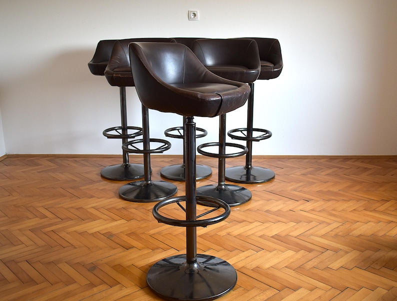 Op de afbeelding: Vijf bruine leren barstoelen met zwarte metalen frames en voetensteunen. De krukken zijn in een halve cirkel op een houten vloer geplaatst.