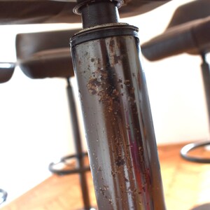 Op de afbeelding: Close-up van een zwarte metalen barstoel met een bruine leren zitting. De stoel heeft een versleten, verweerde afwerking en vertoont tekenen van gebruik.