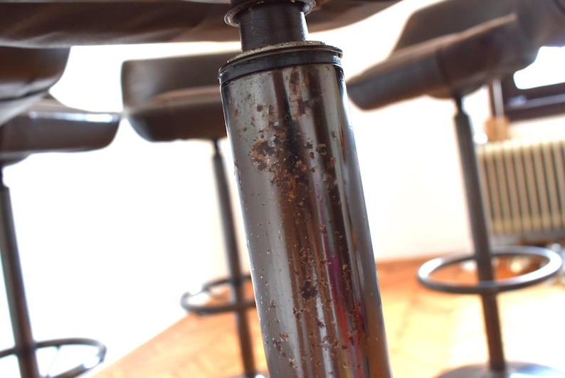 Op de afbeelding: Close-up van een zwarte metalen barstoel met een bruine leren zitting. De stoel heeft een versleten, verweerde afwerking en vertoont tekenen van gebruik.