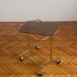 May include: A gold metal serving cart with a smoked glass top and a wire basket underneath. The cart has four wheels and is on a wooden floor.