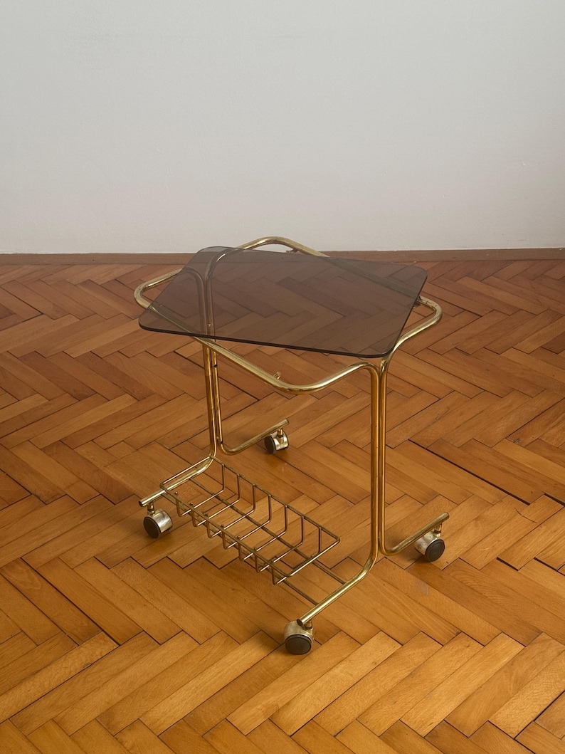 May include: A gold metal serving cart with a smoked glass top and a wire basket underneath. The cart has four wheels and is on a wooden floor.