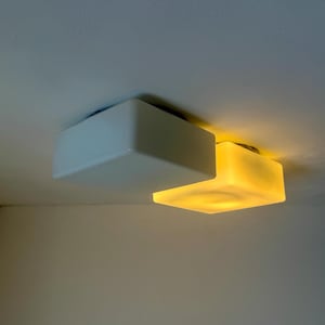 May include: Two white, square ceiling lights are mounted on a white ceiling. One light is off, while the other is illuminated with a warm yellow glow. The lights are modern and minimalist in design.