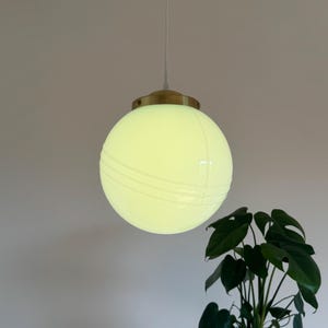 May include: A white globe pendant light with a brass fixture hangs from a white ceiling. The globe has a subtle ribbed texture and emits a warm, yellow light.