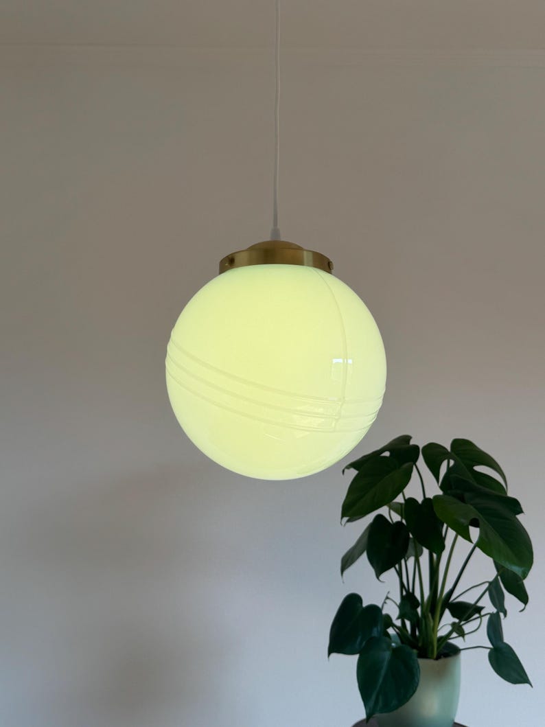 May include: A white globe pendant light with a brass fixture hangs from a white ceiling. The globe has a subtle ribbed texture and emits a warm, yellow light.