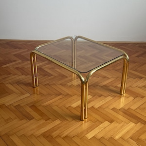 以下が含まれることがあります： 透明なガラス天板と金色の金属フレームの正方形のコーヒーテーブル。 テーブルは、丸みを帯びた角と4本の脚を備えた幾何学的なデザインです。 テーブルの幅は約61cm、高さは41cmです。