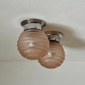 May include: Two flush mount ceiling lights with a chrome base and a pink, ribbed glass shade. The shades are spherical and have a frosted appearance. The lights are mounted on a white ceiling.