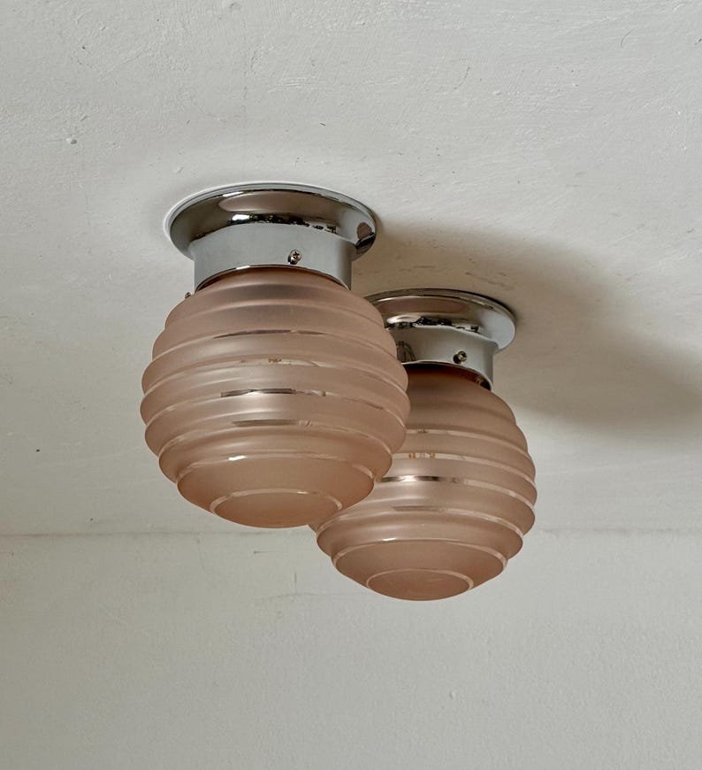 May include: Two flush mount ceiling lights with a chrome base and a pink, ribbed glass shade. The shades are spherical and have a frosted appearance. The lights are mounted on a white ceiling.