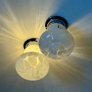 May include: Two flush-mount ceiling lights with chrome bases and globe-shaped, white glass shades. The shades have a speckled pattern. The lights are turned on, casting a warm glow and radiating light patterns on the ceiling.
