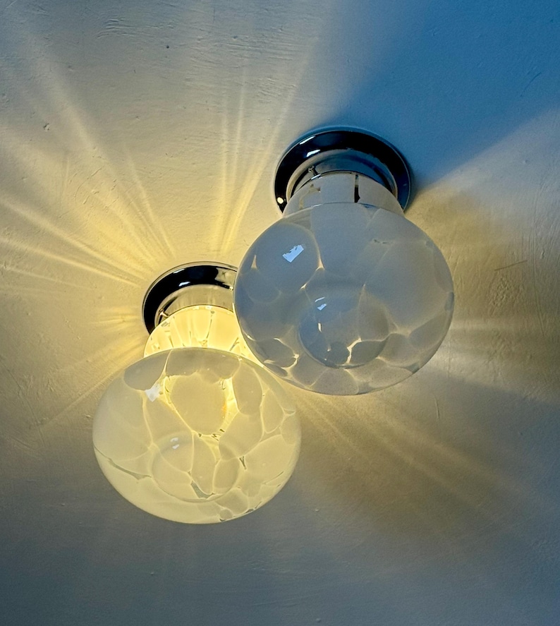 May include: Two flush-mount ceiling lights with chrome bases and globe-shaped, white glass shades. The shades have a speckled pattern. The lights are turned on, casting a warm glow and radiating light patterns on the ceiling.
