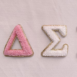 May include: Three Greek letters, Delta Sigma Pi, are embroidered in pink, white, and red with gold trim. The letters are on a white background.