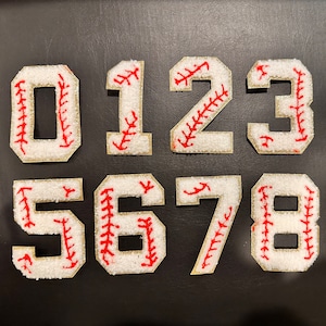 May include: Ten white felt baseball-style numbers with red stitching, from 0 to 9, on a black background.