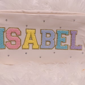 May include: White canvas cosmetic bag with the name "ISABEL" embroidered in colorful letters with gold glitter accents.