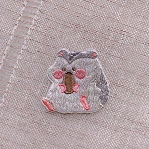 May include: A gray embroidered patch of a hamster holding a brown nut in its paws. The hamster has pink cheeks and a gray hat.