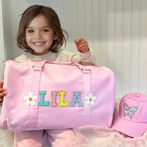 May include: A pink duffel bag with a personalized name "LILA" in colorful letters and two white daisy appliques. A matching pink baseball cap with a butterfly applique is also pictured.