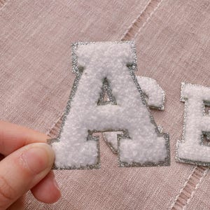 May include: White felt letter A with silver glitter trim. The letter is a patch or applique that can be ironed on or sewn onto fabric.