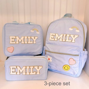 Pu&ograve; includere: Un set di 3 pezzi azzurro chiaro composto da uno zaino, una borsa per il pranzo e un astuccio. Ogni pezzo presenta il nome "EMILY" in bianco con finiture dorate. Il set &egrave; decorato con toppe ricamate di arcobaleni, cuori, fiori e una faccina sorridente.