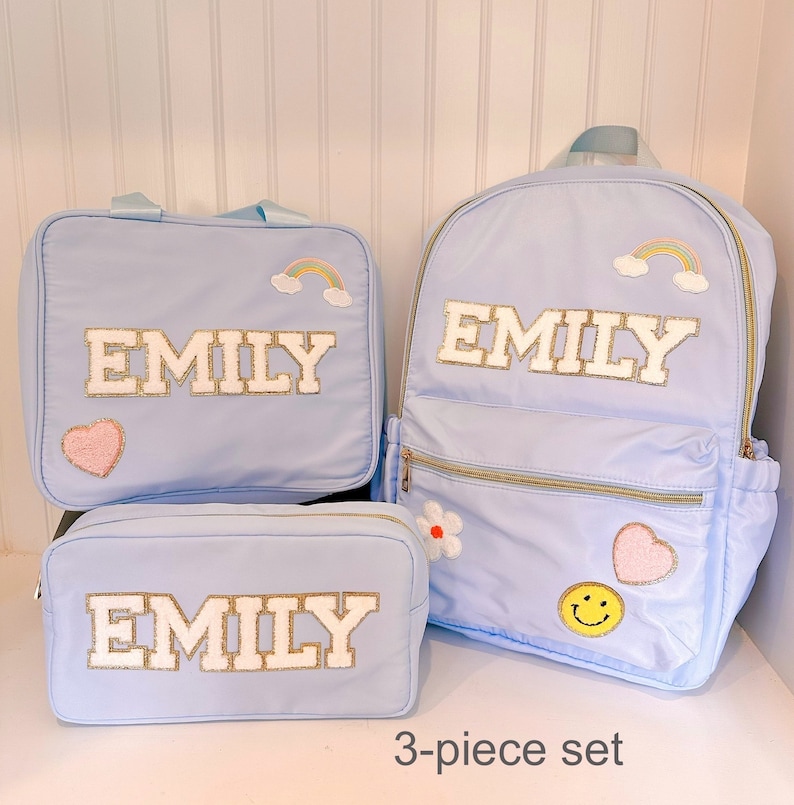 Pu&ograve; includere: Un set di 3 pezzi azzurro chiaro composto da uno zaino, una borsa per il pranzo e un astuccio. Ogni pezzo presenta il nome "EMILY" in bianco con finiture dorate. Il set &egrave; decorato con toppe ricamate di arcobaleni, cuori, fiori e una faccina sorridente.