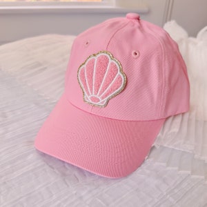 May include: Pink baseball cap with a gold and white seashell patch.