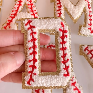 May include: Gold glitter letters with white and red baseball stitching. The letters spell out "GAME".
