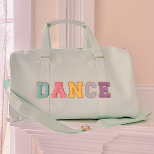 Peut inclure: Un sac de sport vert menthe avec le mot "DANCE" en lettres colorées et texturées. Le sac a une poignée supérieure, une bandoulière et une quincaillerie dorée. Le sac est conçu pour la danse ou la gym.